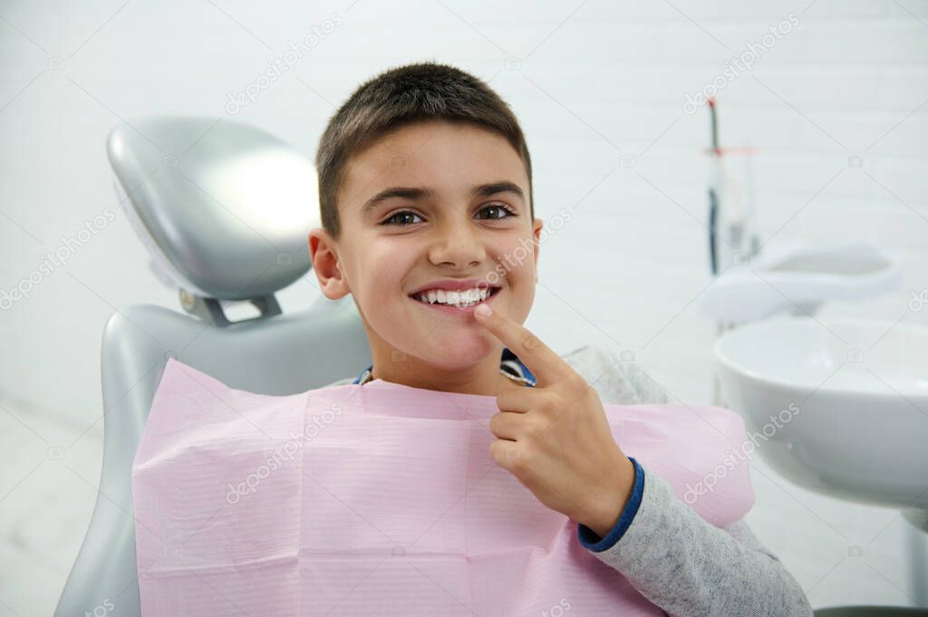 Happy boy holds finger near his mouth, looks at camera, smiles with beautiful toothy smile after receiving dental treatment in dentistry clinic. Oral hygiene, early prevention teeth diseases concept