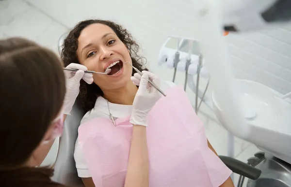 美しい笑顔の若い患者の女性は 衛生士は ステンレス製の歯科用機器を使用して彼女の歯を検査または治療しながら 彼女の口を開いて歯科医の椅子に座っている 鏡とプローブ — ストック写真