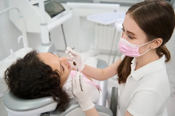 ピンクの医療保護マスクの歯科衛生士は 計画された医療検査中に歯科医院で滅菌ステンレス製の歯科用鏡とプローブを持つ女性患者の口腔を調べる — ストック写真