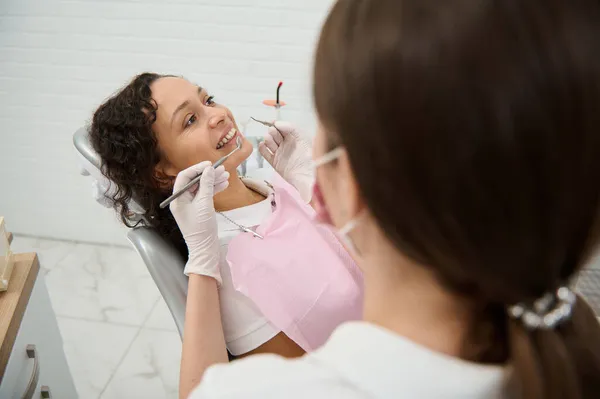 ステンレス製の歯科用器具を持つぼやけた歯科衛生士を通して眺め 歯医者の椅子に座って完璧な白い歯を持つ歯科クリニックの女性患者の口腔を調べる — ストック写真