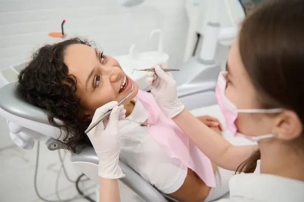 歯科クリニックで歯科検査中の歯科医の椅子の美しい女性 歯科衛生士がステンレス製の歯科用器具を持ち 前景に患者の口腔を検査 — ストック写真