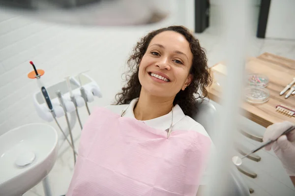 Güzel dişlek gülüşlü, dişçi koltuğunda oturan ve modern dişçilik kliniğinde diş muayenesini bekleyen güzel bir Afrikalı kadının yüksek açılı görüntüsü. Ağız bakımı, diş temizliği konsepti