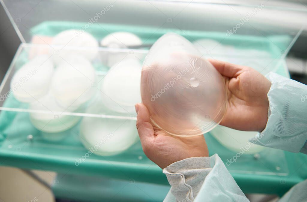 Breast prothesis in surgeon hands on background of medical samples of breast silicone implants of various sizes for breast augmentation aesthetic plastic surgery after resection due to cancer disease
