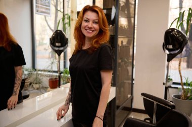 Kızıl saçlı çekici kadın kuaför siyah üniformalı kuaför kuaför güzellik salonunda aynanın yanında duruyor, dişlek gülümsemesiyle müşteriyi beklerken kameraya bakıyor.