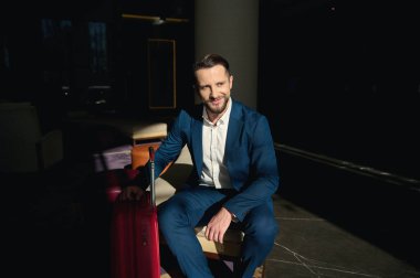 Yakışıklı bir iş adamı, başarılı bir girişimci günlük takım elbiseli, yatırım danışmanı otel lobisindeki bir sandalyeye oturuyor ve gülümsüyor, iş seyahatinde yan tarafa bakıyor.