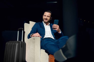 Yakışıklı genç iş adamı, yatırımcı, girişimci, bir otel lobisinde oturan, elinde cep telefonu, bacak bacak üstüne atmış, gülümseyen, iş seyahatinde yan tarafa bakan..