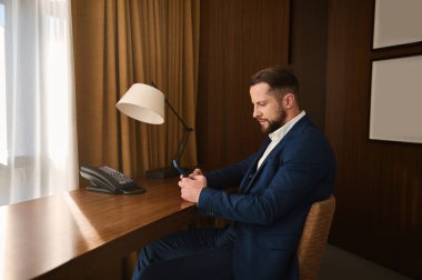 Başarılı genç bir iş adamı, yakışıklı girişimci, koyu renk takım elbiseli yatırımcı otel odasında bir masada oturuyor ve cep telefonu kullanıyor, iş seyahatleri sırasında göz gezdirme sohbeti yapıyor.