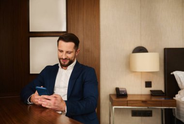 Yakışıklı, varlıklı bir iş adamının özgüvenli portresi başarılı bir iş adamı, girişimci, otel odasında dinlenen koyu takım elbiseli yatırımcı, iş gezisi sırasında cep telefonu kullanırken gülümseyen