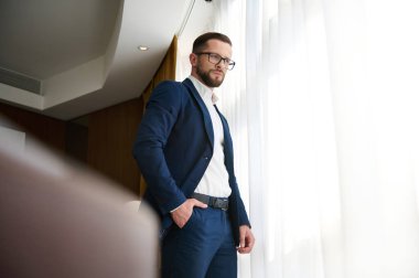 Yakışıklı bir iş adamı, başarılı bir yatırımcı, takım elbiseli bir girişimci, bir iş gezisi sırasında bir otel odasının penceresinden şehir manzarasına bakıyor.