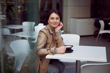 Güzel, çekici, çekici esmer, kısa saçlı, bej renkli trençkotlu, sokak kafeteryasında bir masada oturan ve rüya gibi görünümlü bir sabah kahvesinden hoşlanıyor.