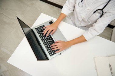Beyaz klinikteki tıp bürosunda dizüstü bilgisayarda yazı yazan kadın doktorların yüksek açılı görüntüsü.