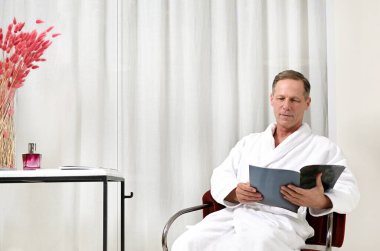 Beyaz bornozlu yakışıklı Avrupalı bir adam, bir koltukta oturup dergi okuyor ve erkek güzellik tedavisi gördükten sonra özel sağlık merkezinin spa odasında dinleniyor.