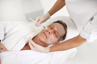 Tanımlanamayan bir güzellik uzmanının yaşlanmayı önleyen spa tedavisinde olgun bir adamın yüzüne bir fırçayla nemlendirici yüz maskesi uygulaması. Boşluğu kopyala