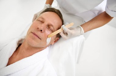 Orta yaşlı yakışıklı bir adamın tepeden bakışı. Gözleri kapalı. Yüzünde erkek güzellik tedavileri görüyor. Tanınmayan kozmetik uzmanı nemlendirici kozmetik ürünlerini ahşap bir spatulayla uyguluyor.