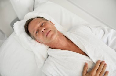 Yakışıklı, rahat, olgun, 50 yaşında beyaz bornozlu Avrupalı bir adamın beyaz lüks kaplıca kliniğinde masaj masasında yatarken kameraya baktığı yüksek açı.