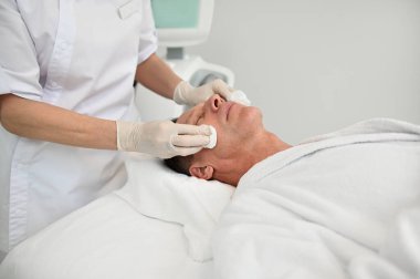 Kaplıca salonundaki olgun bir erkek müşteriye yakın çekim. Profesyonel güzellik tedavisi görüyor. Pamuk pedlerle tanınmaz hale gelen kadın güzellik uzmanı. Deri bakımı konsepti. Erkeksi güzellik.