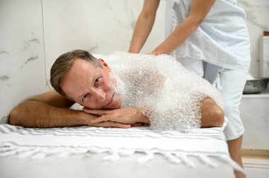 Yakışıklı, orta yaşlı, Türk hamamında köpük banyosu yapan, kameraya bakan, mermer masaya uzanmış, tanınmayan bir masözün sağlık merkezinde köpük masajı yaptığı bir adam.