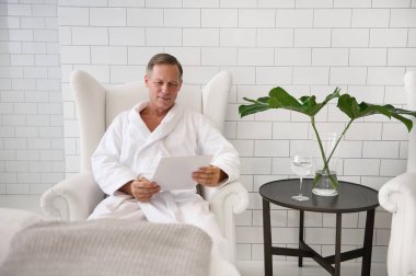 Yakışıklı, orta yaşlı, 50 yaşında, beyaz bornozlu bir iş adamı elinde bir bardak limon suyu, gülümsüyor, dergi okuyor ve özel sağlık merkezinde dinleniyor.