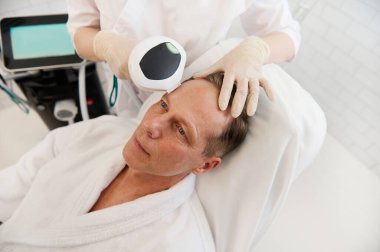 Yakışıklı, olgun bir adam, yüz yenileme, yüz gerdirme ve kozmetik kliniğinde fizyoterapi için benimle yüz hatları oluşturma prosedürü uyguluyor. Profesyonel yaşlanma karşıtı terapi.