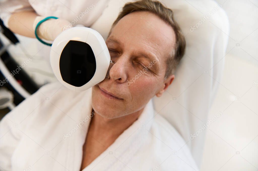 Close-up shot for advertisement of a handsome European man relaxing in wellness spa during mesotherapy course with modern medical equipment for face tightening at cosmetology clinic