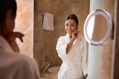 Çekici esmer, beyaz bornozlu orta yaşlı Avrupalı kadın mermer bir banyoda dikiliyor ve aynadaki yansımasına gülümsüyor..