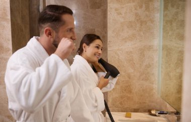 Birbirine aşık genç, güzel heteroseksüel bir çiftin portresi. Beyaz terli bornozlar giyiyorlar. Dişlerini fırçalıyorlar ve saçlarını kurutuyorlar. Mutlu sabah hijyen rutin konsepti