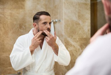 Beyaz bornozlu genç yakışıklı Avrupalı adam banyodaki aynadaki yansımasına bakıp sabah temizliği sırasında sivilcelerini ve sivilcelerini kontrol ediyor.