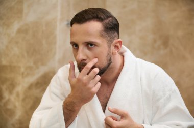 Beyaz bornozlu genç bir adamın aynadaki yansıması görüntüsünden endişe duyuyor, banyoda kendine bakıyor ve sabah temizliği sırasında yüzünü sivilce için kontrol ediyor.