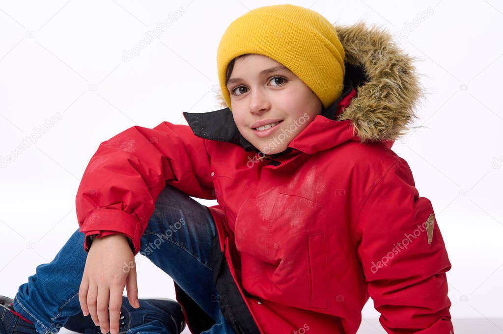 Beautiful child, handsome preadolescent European boy in bright yellow hat and warm red down jacket smiles toothy smile looking at camera sitting on a white background with copy space for ads