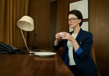 Genç, güzel bir iş kadını, girişimci, göz ve iş kıyafetlerinde başarılı bir satış müdürü iş gezisi sırasında otel odasında dizüstü bilgisayarla oturup kahve fincanı tutuyor.