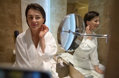 Güzel bakımlı esmer, beyaz bornozlu çekici genç bir kadın lüks bir banyoda dikiliyor ve aynadaki yansımasına bakıyor, kendisine hayran. Cilt bakımı kavramı