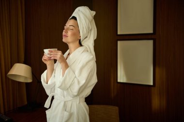 Beyaz terli havlulara sarılı keyifli genç bir kadın, banyodan sonra gözleri kapalı duruyor, bir fincan kahve tutuyor, iş gezisi sırasında bir otel odasında yalnızlık ve huzur içinde vakit geçiriyor.