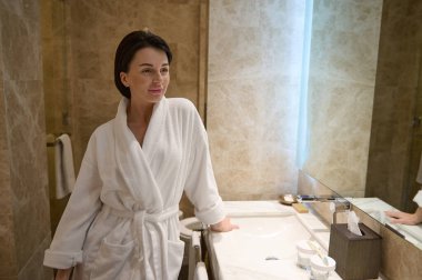 Göz kamaştırıcı genç esmer, beyaz bornozlu güzel kadın mermer duvarlı lüks bir banyoda aynanın yanında duruyor. Sabah hijyen programı ve cilt bakımı konsepti