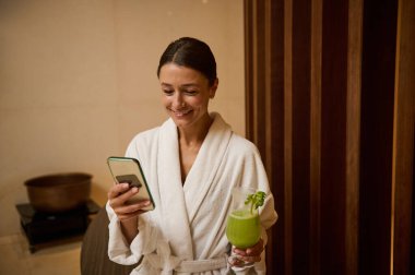 Çekici, orta yaşlı, beyaz terry bornozlu bir kadın. Elinde sağlıklı taze sıkılmış vitamin meyve suyu var. Sağlık merkezindeki dinlenme yerinde cep telefonu kullanıyor.