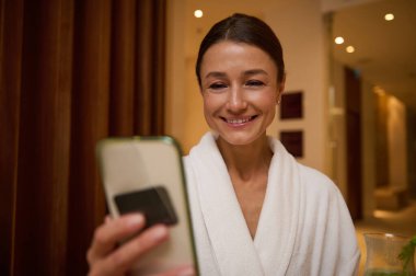 Beyaz bornoz giymiş, orta yaşlı beyaz bir kadına odaklan. Parlak sağlık merkezlerinde dinlenirken dişlek bir gülümsemeyle cep telefonu kullan.