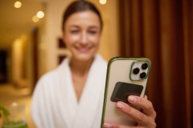 Bulanık, bulanık, odaktan yoksun, oldukça koyu renk saçlı bir kadının elindeki cep telefonuna odaklanın. Sağlık merkezindeki spa merkezinde dinlenen beyaz terli kumaş bornoz giyin.
