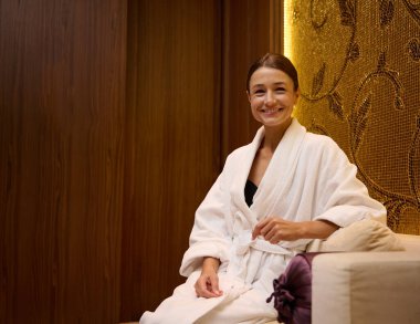 Rahat, orta yaşlı, beyaz bornozlu güzel bir kadının rahat bir orta yaşlı portresi kanepede oturuyor, dişlek bir gülümseme ile kameraya bakıyor ve lüks bir sağlık spa salonunda dinleniyor.