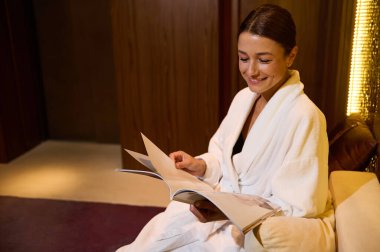 Orta yaşlı zarif bir kadın, beyaz terry bornoz giyiyor, dişlek gülüşü takınıyor, sağlık merkezinde dinlenirken dergi okuyor. Uzayı kopyala. Kozmetoloji, vücut bakımı konsepti