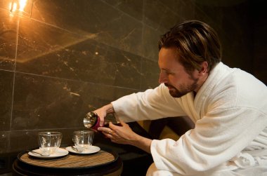 Rahatlamış, orta yaşlı, Avrupalı bir adam spa salonunda dinleniyor, beyaz bir bornozla güneşleniyor ve şeffaf cam sürahisinden bardaklara bitki çayı döküyor.