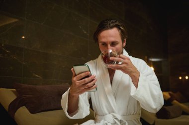 Beyaz atlı beyaz bornozlu çekici, rahat beyaz adam akıllı telefon kullanıyor, sağlıklı bitkisel içecekler içiyor, havuz başında yalnızlığın ve huzur verici spa merkezinin tadını çıkarıyor.