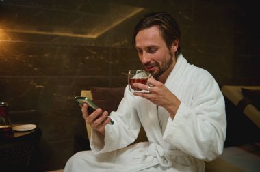 Rahat, çekici, orta yaşlı, beyaz, bornozlu, çay içen, elinde akıllı bir telefona bakan, göz gezdiren, jakuzinin yanında güzellik merkezinde dinlenen,