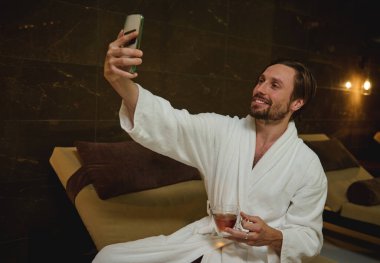 Bornozlu yakışıklı genç Avrupalı bir adam sağlık spa merkezinde jakuzinin yanında oturuyor, elinde bitki çayı tutuyor ve gülümsüyor dişlek bir gülümseme ile özçekim yapıyor ya da spa merkezinde dinlenirken video aracılığıyla konuşuyor.