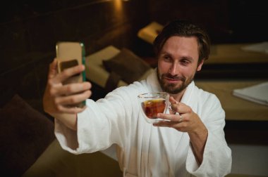 Yakışıklı beyaz adam, dinlenme odasında jakuzinin yanında oturuyor, çay içiyor ve uzanmış elinde kamerayla kameraya bakıyor, selfie çekiyor, lüks spa merkezlerinde tatillerin tadını çıkarıyor.