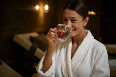 Beyaz bornozlu zarif, orta yaşlı Avrupalı kadın, sıcak, sağlıklı bitkisel çay içen, jakuzinin yanındaki şezlongda oturan, spa salonundaki spa prosedürlerinin tadını çıkaran.