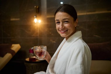 Beyaz bornozlu zarif beyaz kadın, elinde bir bardak sıcak, sağlıklı bitkisel içecek, kameraya bakıyor, jakuzinin yanındaki şezlongda oturuyor, spa salonundaki spa prosedürlerinin tadını çıkarıyor.