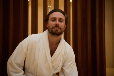 Çekici, orta yaşlı, çekici bakışlı, tıraşsız, beyaz bornozlu yakışıklı, maço bir adam lüks bir spa merkezinin lobisinde durmuş, kendine güvenen ve gizemli bir şekilde kameraya bakıyor.