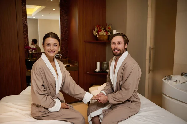 Mutlu orta yaşlı aşık heteroseksüel çift, bornoz giyiyorlar, el ele tutuşuyorlar, masaj masalarına oturuyorlar ve gülümsüyorlar, kameraya bakıyorlar. Yeni evliler balayına sağlık merkezlerinde çıkıyorlar.