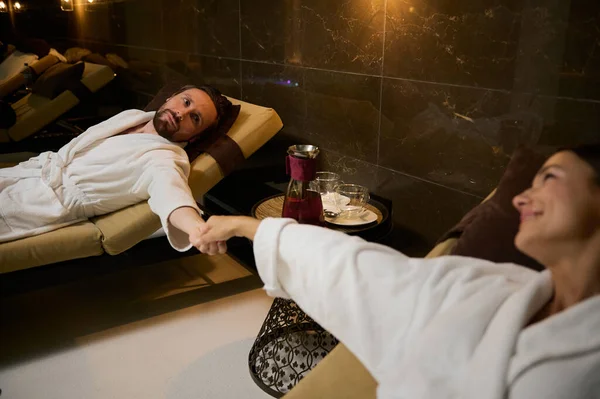 Yeni evli, güzel bir çift beyaz bornozlu yakışıklı bir adam güneş pansiyonunda uzanıyor ve sevgilisinin elini tutarken sağlık merkezindeki balayının tadını çıkarıyor.