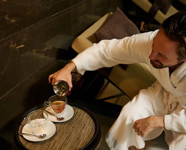 Rahatlamış, orta yaşlı, beyaz bornozlu, güneşli bir odada dinlenen ve şeffaf cam sürahisinden bardaklara bitki çayı döken beyaz tenli, orta yaşlı, beyaz bir adamın manzarası.