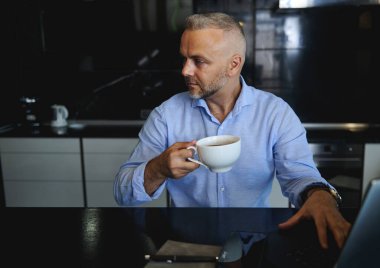 Çekici, orta yaşlı, günlük giysiler içinde beyaz bir adam. Sabah kahvesinin keyfini çıkarıyor. Dizüstü bilgisayarıyla modern, yüksek teknolojili bir mutfağın arka planında oturuyor.